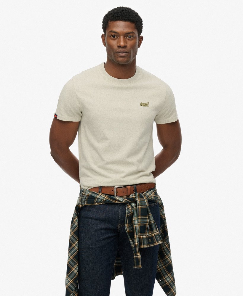 Man wearing a beige t-shirt with a logo, plaid shirt over shoulders, and dark jeans on a white background