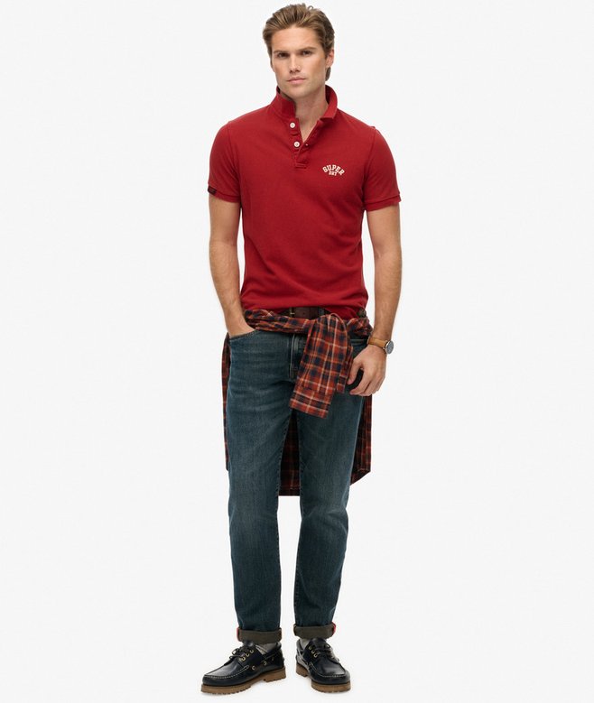 Man wearing a red polo shirt, plaid shirt over shoulders, and jeans on a white background