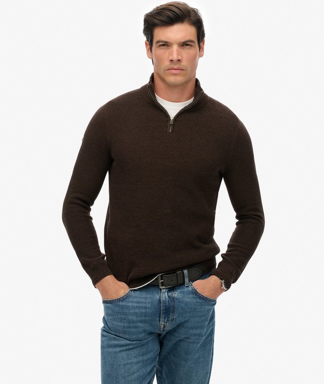 Man wearing a brown quarter-zip sweater and blue jeans on a white background