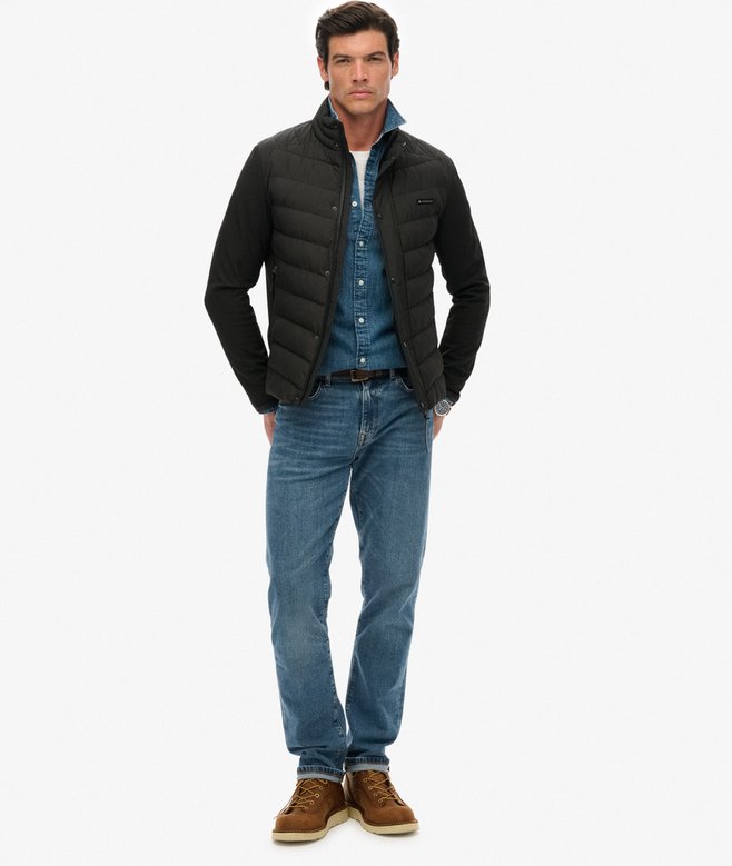 Man wearing a black puffer jacket, blue shirt, jeans, and brown shoes on a white background