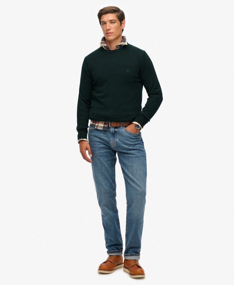 Man wearing a dark green sweater, plaid shirt, blue jeans, and brown shoes on a white background