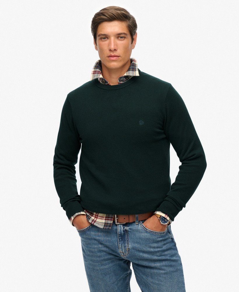 Man wearing a dark green sweater over a plaid shirt with blue jeans on a white background