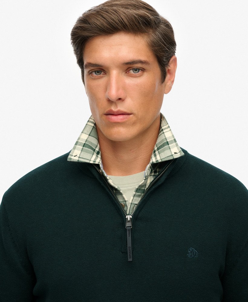 Man wearing a dark green sweater over a plaid shirt on a light gray background
