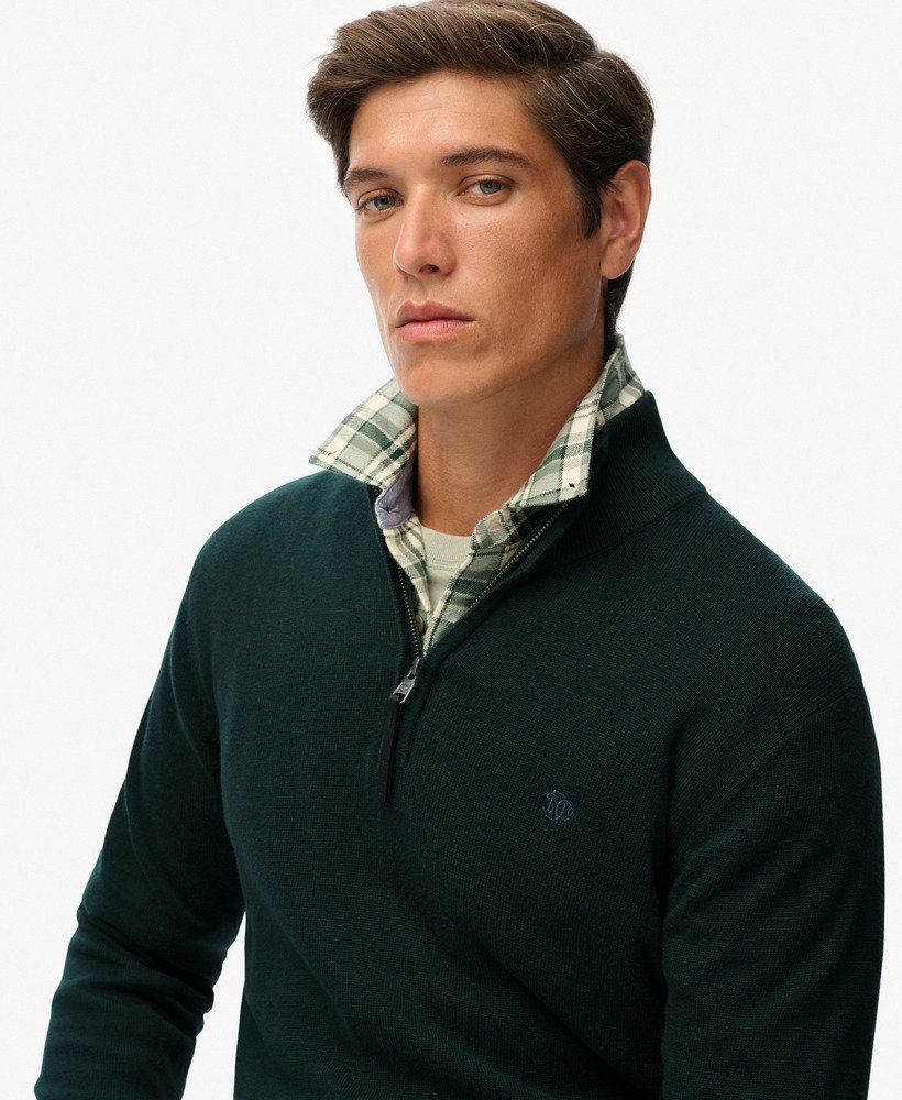Man wearing a dark green sweater over a plaid shirt on a white background
