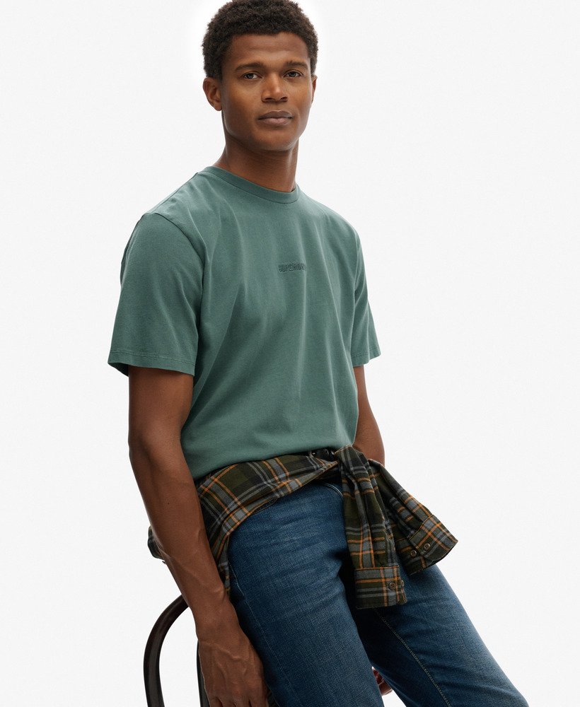 Man wearing a green t-shirt and plaid shirt over jeans, sitting on a chair with a white background