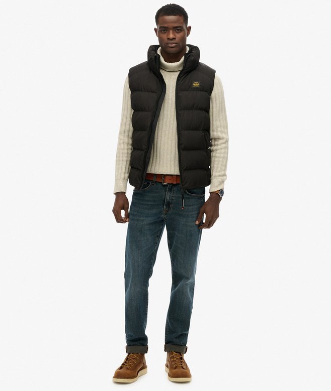 Man wearing a black puffer vest over a sweater, jeans, and boots on a white background