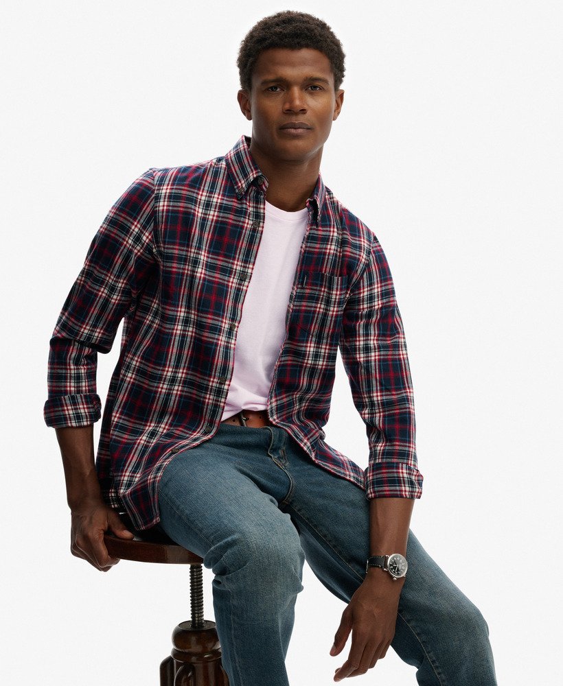 Man wearing a plaid shirt over a white t-shirt and jeans, sitting on a chair with a plain background