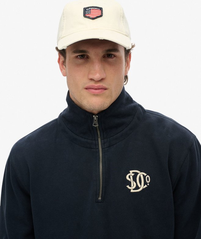 Person wearing a navy blue quarter-zip sweatshirt with a logo and a beige cap with an American flag emblem on a white background.