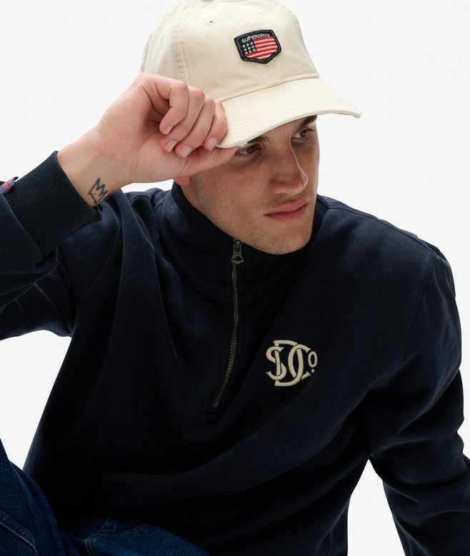 Person wearing a navy sweatshirt with a logo and a beige cap on a white background