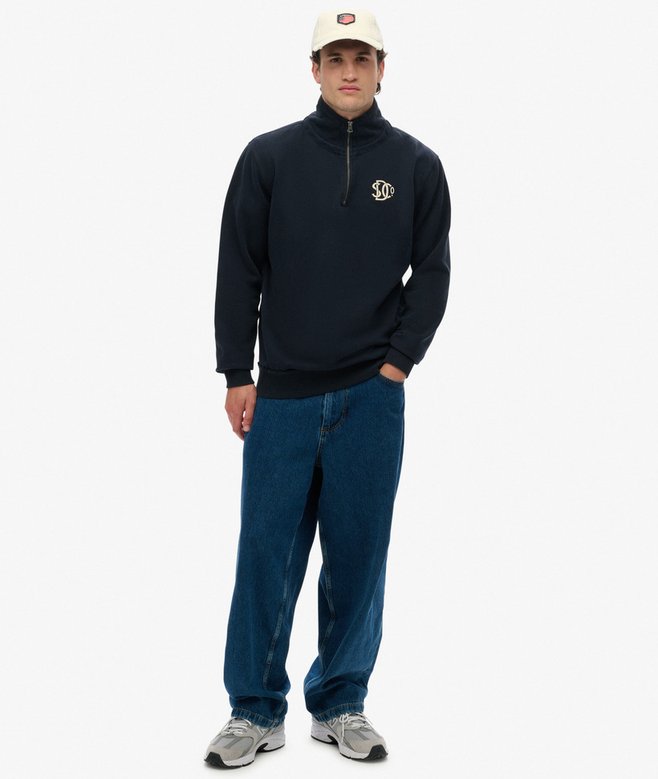 Man wearing a navy blue sweater with a logo, blue jeans, and a white cap on a white background