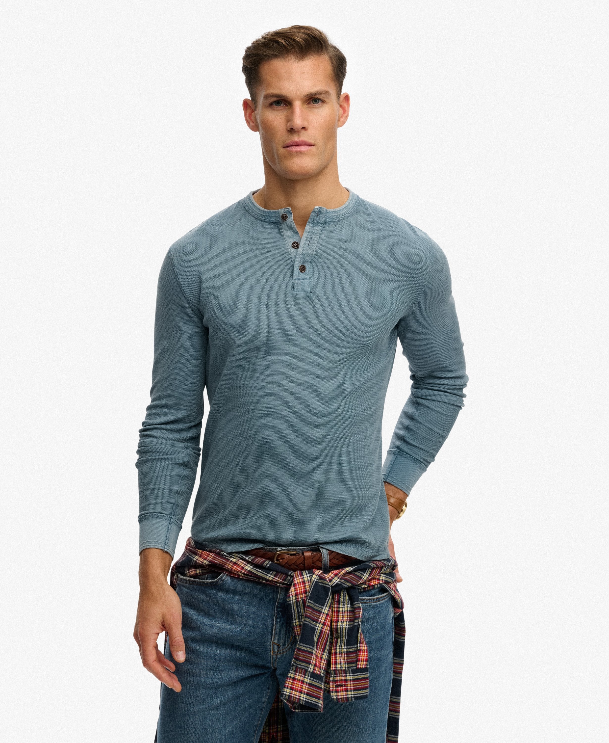 Man wearing a blue long-sleeve henley shirt and plaid pants on a white background