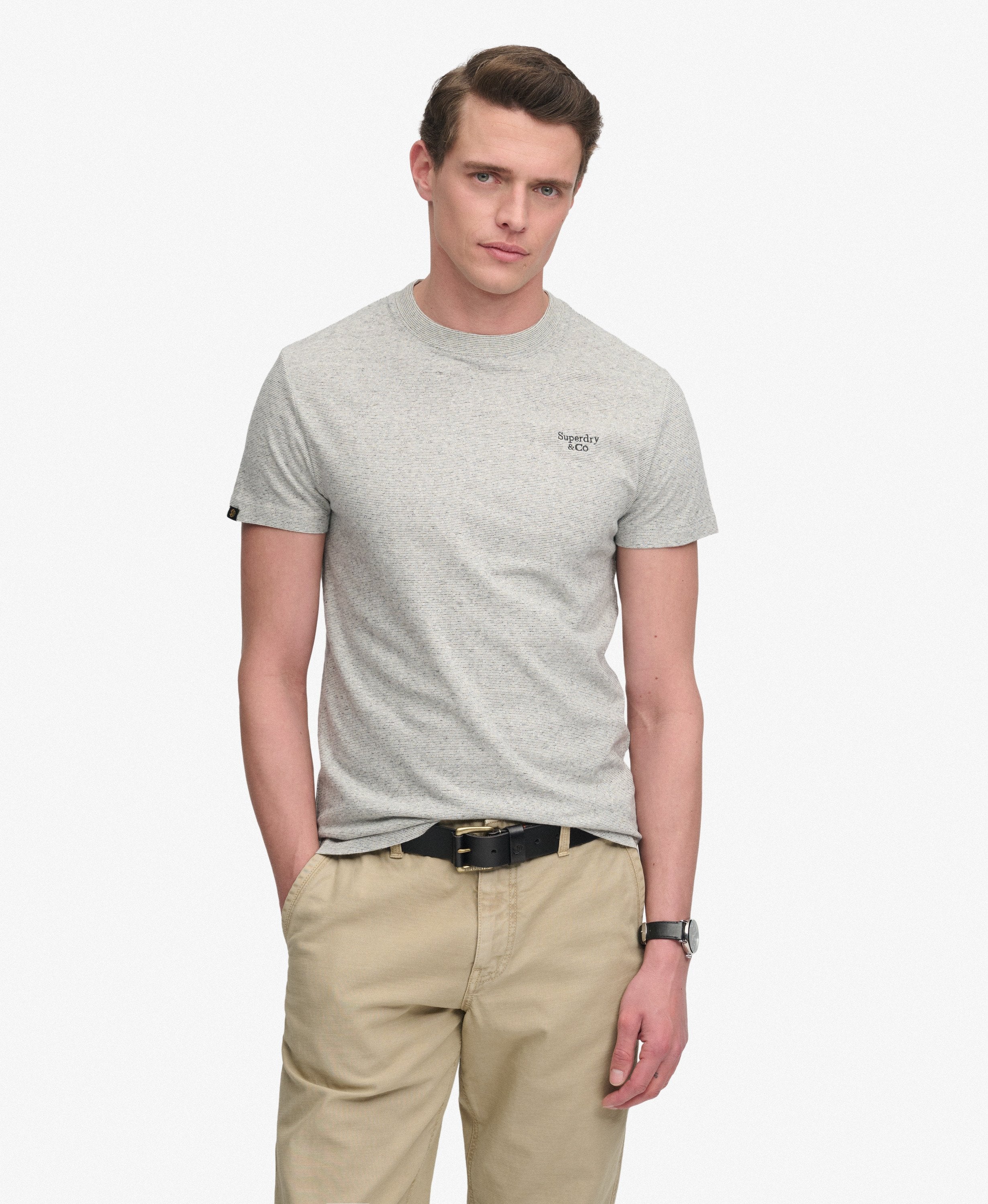 A man wearing a light gray heather crew-neck t-shirt with a small black embroidered logo on the left chest, paired with khaki pants and a black belt, against a plain light gray background.